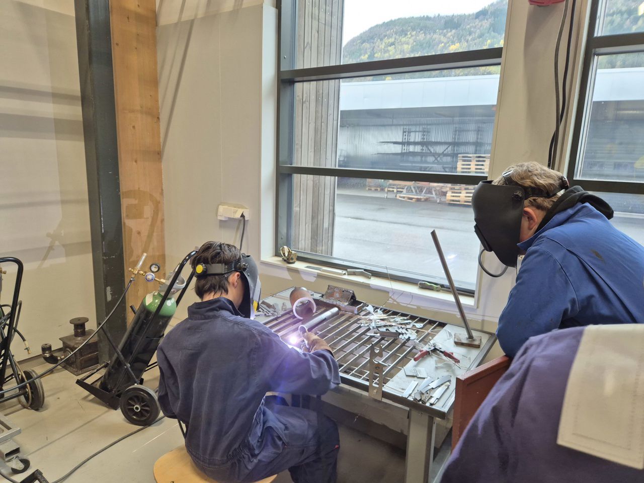 to ungdommar i kjeledress har på sveisemaske. Ein av ungdommane sveisar medan den andre ser på