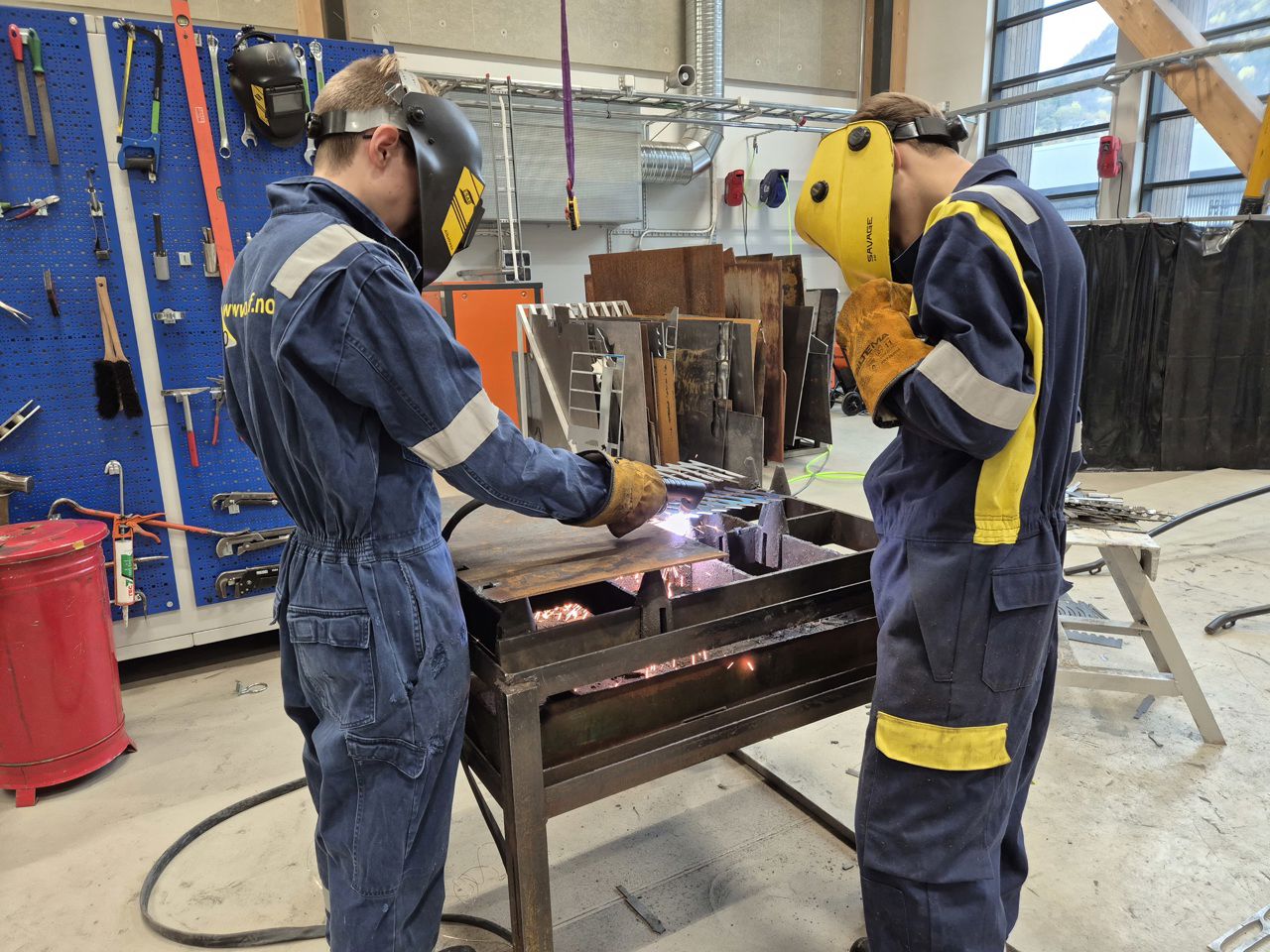 to ungdommar i blå kjeledress står med sveisemaske og hanskar. Ein av ungdommane står og sveisar og den andre står og ser på. 