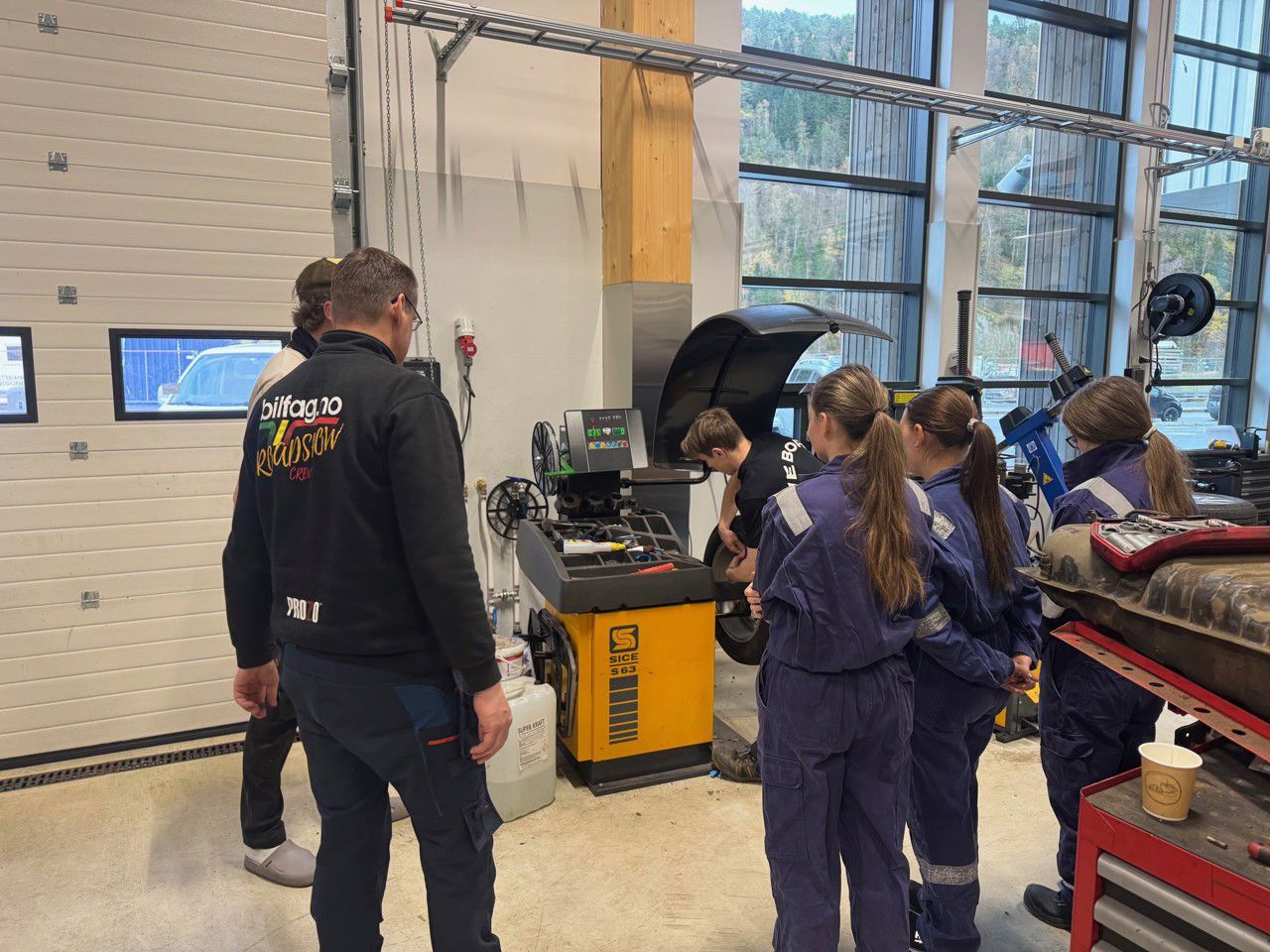 tre jenter med blå kjeledress står sammen med to vaksne og ser på ein gut som står og jobbar med eit hjul. 