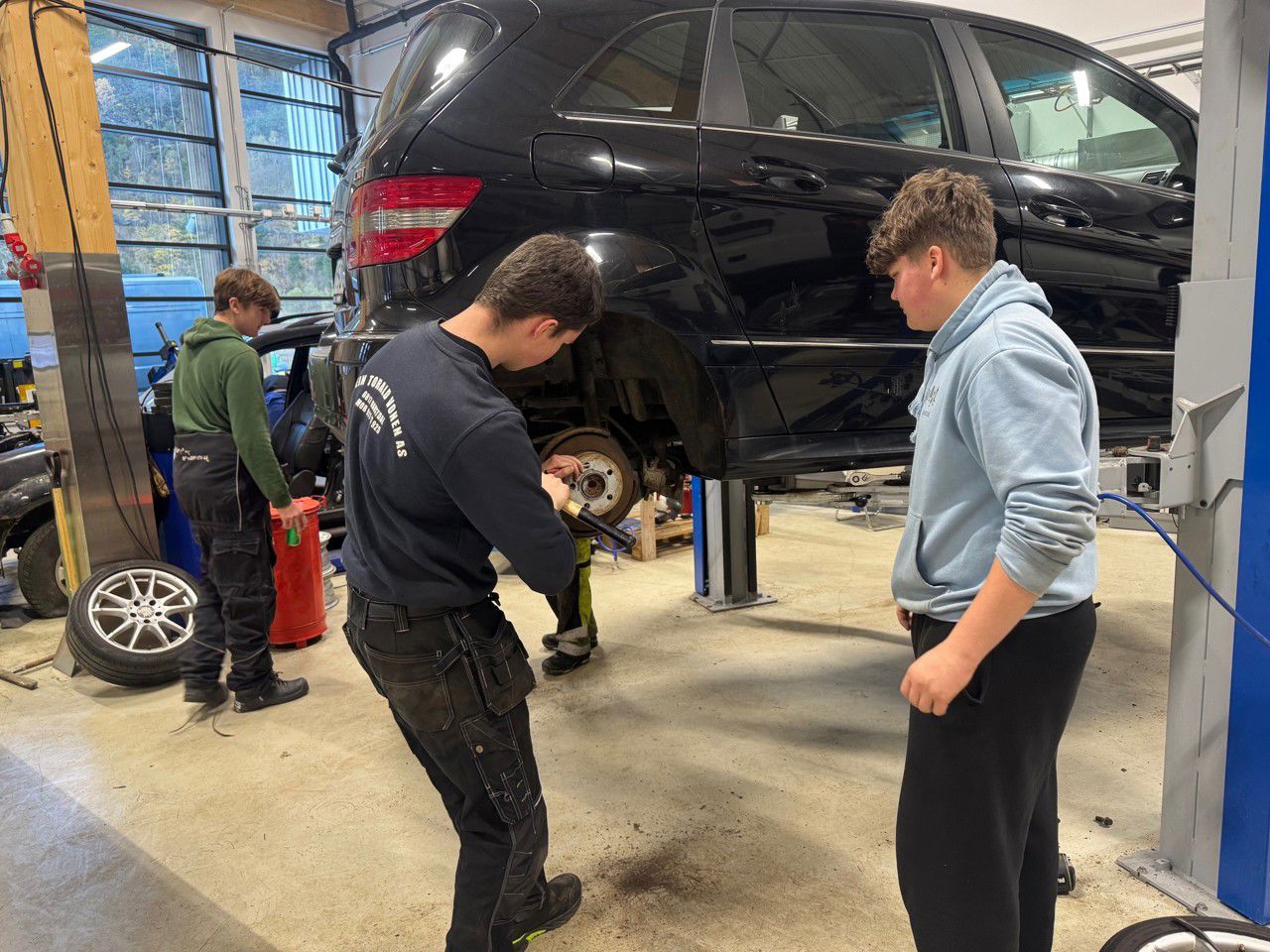 Ein gut står og jobbar ved høgre bakaksling på ein bil, som er sett opp i arbeidshøgd. Ein annan gut står og ser på arbeidet. Ein tredje gut står bak bilen.  