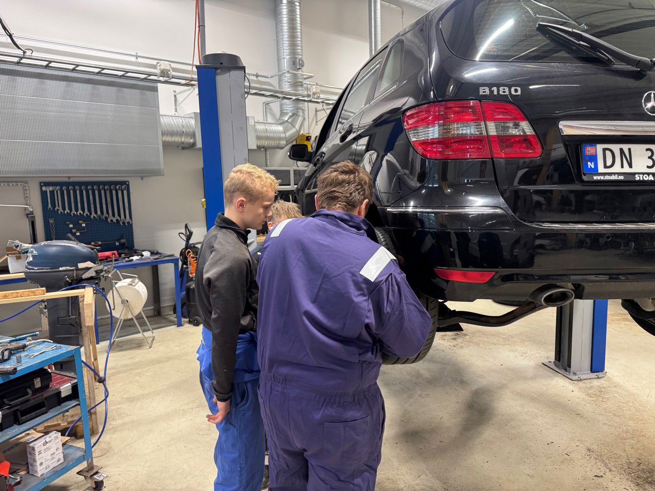 Ein gut i blå kjeledress står saman med to andre guar og jobbar på ein bil ved venstre bakaksling. Bilen er heva opp i arbeidshøgd. 